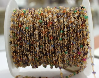 May include: A spool of beaded chain featuring small, multi-coloured gemstone beads strung on gold-coloured wire. The beads are various colours, including purple, green, orange, and clear. The chain is wound around a white plastic spool.