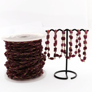 May include: A spool of ruby red gemstone chain with gold-toned metal links. The chain is displayed on a black stand with several strands of the chain hanging down.