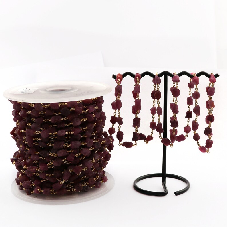 May include: A spool of ruby red gemstone chain with gold-toned metal links. The chain is displayed on a black stand with several strands of the chain hanging down.