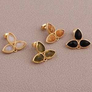 Puede incluir: Cuatro pares de pendientes de botón con un engaste dorado. Cada pendiente presenta tres piedras en forma de lágrima en varios colores: blanco, marrón, verde y negro. Los pendientes se muestran sobre un fondo neutro.