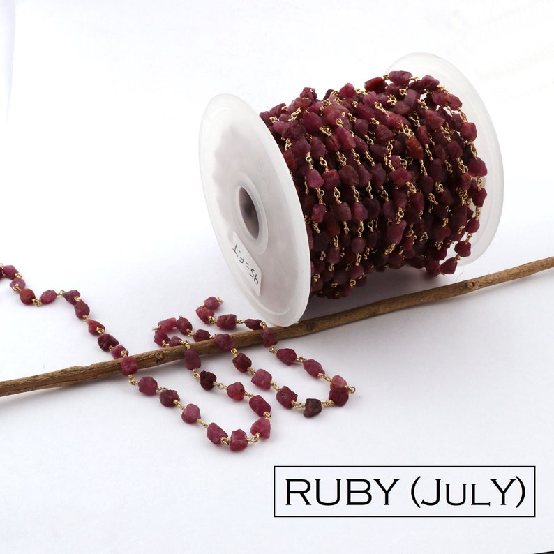 May include: A spool of gold chain with ruby beads. The chain is made of small, irregular ruby beads that are strung together with gold wire. The chain is wound around a white spool with the text "HS-FT" printed on it. The chain is lying on a wooden stick.