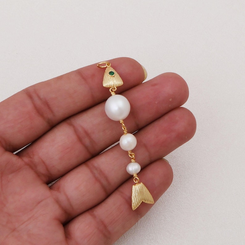 May include: A gold-toned pendant shaped like a fish, featuring a textured triangular head with a small green stone eye, three white spherical pearls of varying sizes forming the body, and a textured triangular tail piece at the bottom.