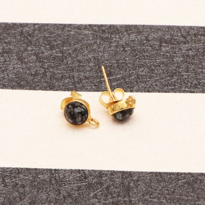 May include: A pair of gold-toned stud earrings featuring a circular black and grey mottled stone inlay. Each earring has a small metal loop attached to the side of the setting and includes a matching gold-toned butterfly earring back.