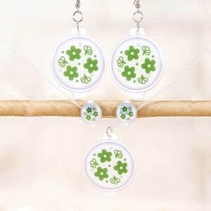 Puede incluir: Un collar, pendientes y un colgante blancos con un diseño floral verde. El collar y los pendientes tienen una cadena plateada.