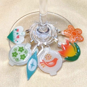 May include: Set of six colorful glass wine glass charms with different designs, including a heart, a flower, a bird, a star, and two floral patterns. The charms are attached to silver rings.