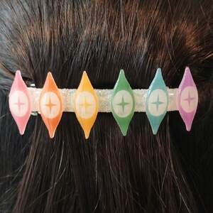 May include: A rainbow-colored hair clip with a white glitter base and a star design on each of the seven teardrop-shaped pieces.