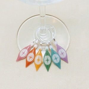 May include: Six colorful glass wine charms with a star design. The charms are hanging from a wine glass.