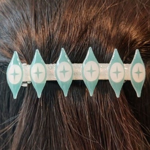 May include: A teal blue hair clip with a silver bar and six white stars. The stars are inside of a diamond shape.