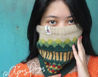 May include: A knitted neck warmer featuring a beige ribbed top section, a green diamond pattern, a mustard yellow vertical stripe band, and a dark green base with small red accents. A small white fabric tag with a graphic is attached to the top edge.