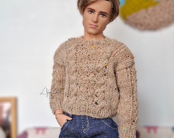 May include: A doll with brown hair wearing a beige cable-knit sweater with small multicolored flecks and dark blue denim jeans. The sweater features a textured pattern with vertical cables and long sleeves. The doll has a neutral facial expression.