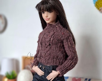 May include: A doll with long brown hair wearing a dark brown cable-knit sweater with a high neck and long sleeves, paired with dark blue denim shorts featuring a frayed hem. The sweater has a textured pattern and subtle multi-colored flecks.