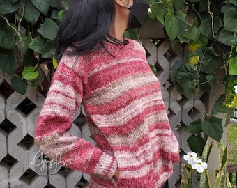 May include: A long-sleeved knit sweater featuring horizontal stripes in shades of deep red, soft pink, and muted beige. The garment has a round neckline, ribbed cuffs, and a ribbed hem, with a visible pocket opening on the lower front side.