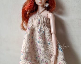 May include: A doll with long red hair wearing a beige knit halter top featuring small blue and pink speckles, paired with blue denim pants. The doll has a pale complexion and articulated limbs. A signature is visible on the right side.
