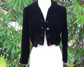 May include: A black velvet cropped jacket with a rounded collar and three matching buttons. The long-sleeved jacket is displayed on a white mannequin against a backdrop of green foliage. The jacket's fabric has a soft, luxurious appearance.