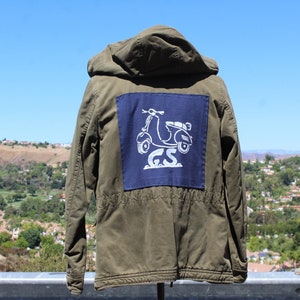 May include: An olive green hooded jacket with a navy blue patch on the back. The patch features a white scooter graphic and the letters "GS". The jacket is made of a durable material and has a drawstring waist.