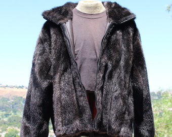 May include: A dark brown faux fur jacket with a zip-up front and a collared neckline. The jacket is displayed on a mannequin, revealing a brown shirt underneath. The background features a clear blue sky and a distant landscape.
