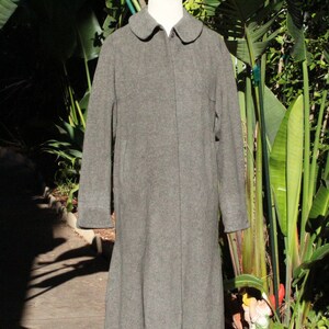 May include: A gray wool coat with a white collar. The coat has long sleeves and a flared skirt. The coat is displayed on a mannequin in front of a lush green background.