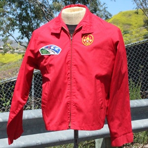 May include: A red jacket with a zip fastening and a collared neckline. The jacket has two patches: one with a blue and white design and another with a gold emblem. The jacket is displayed on a mannequin.