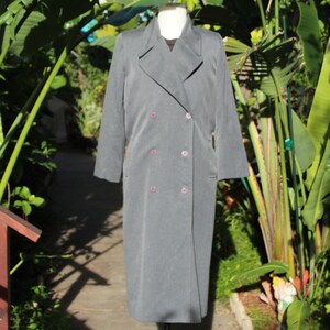 May include: A grey, double-breasted coat with a long length, featuring a notched collar and six pink buttons. The coat is displayed on a mannequin, set against a backdrop of lush green foliage and a wooden railing.