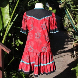 May include: A red two-piece outfit with a floral pattern. The top has a black yoke with turquoise and white trim. The skirt has a matching pattern with turquoise and black trim. The outfit is displayed on a mannequin.