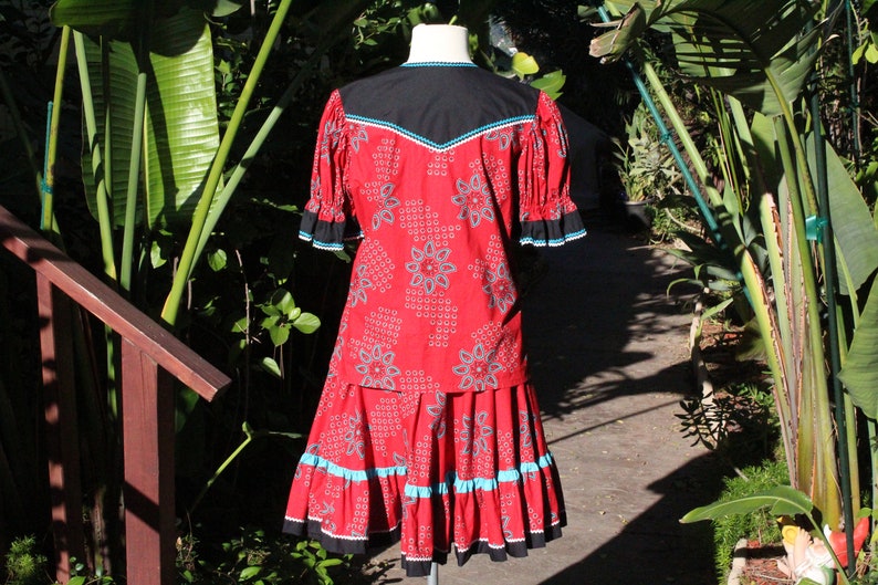 May include: A red two-piece outfit with a floral pattern. The top has a black yoke with turquoise and white trim. The skirt has a matching pattern with turquoise and black trim. The outfit is displayed on a mannequin.