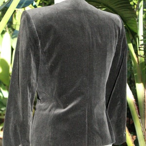 May include: A black velvet blazer with long sleeves. The back view of the jacket shows a tailored fit, with a subtle sheen. The blazer is displayed on a white mannequin form, set against a backdrop of green foliage.