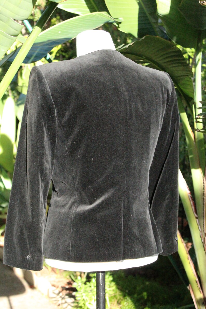 May include: A black velvet blazer with long sleeves. The back view of the jacket shows a tailored fit, with a subtle sheen. The blazer is displayed on a white mannequin form, set against a backdrop of green foliage.