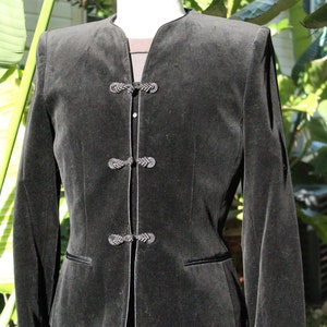 May include: A black velvet jacket with a mandarin collar and decorative frog closures. The jacket has long sleeves and a tailored fit, with a contrasting trim along the edges and pockets. The garment is displayed on a mannequin.