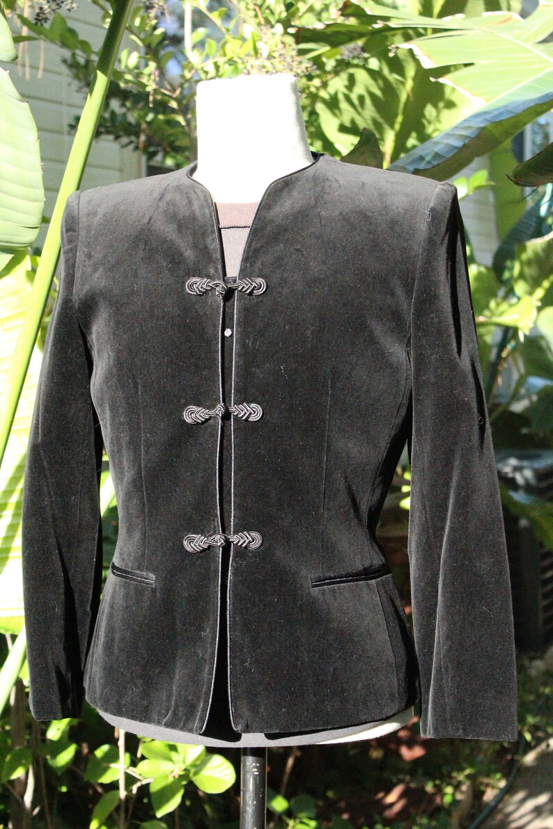 May include: A black velvet jacket with a mandarin collar and decorative frog closures. The jacket has long sleeves and a tailored fit, with a contrasting trim along the edges and pockets. The garment is displayed on a mannequin.