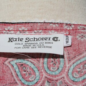 May include: Close-up of a red paisley patterned fabric with a white label that reads "Kate Schorer C." and "Made in U.S.A.". The label also indicates the size is medium.