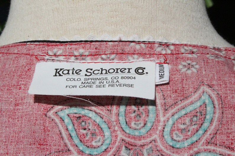 May include: Close-up of a red paisley patterned fabric with a white label that reads "Kate Schorer C." and "Made in U.S.A.". The label also indicates the size is medium.