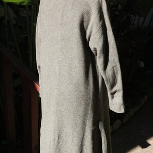 May include: A long, gray wool coat with a wide collar and long sleeves. The coat has a slight A-line shape and a split in the back for ease of movement. The coat is displayed on a white mannequin.