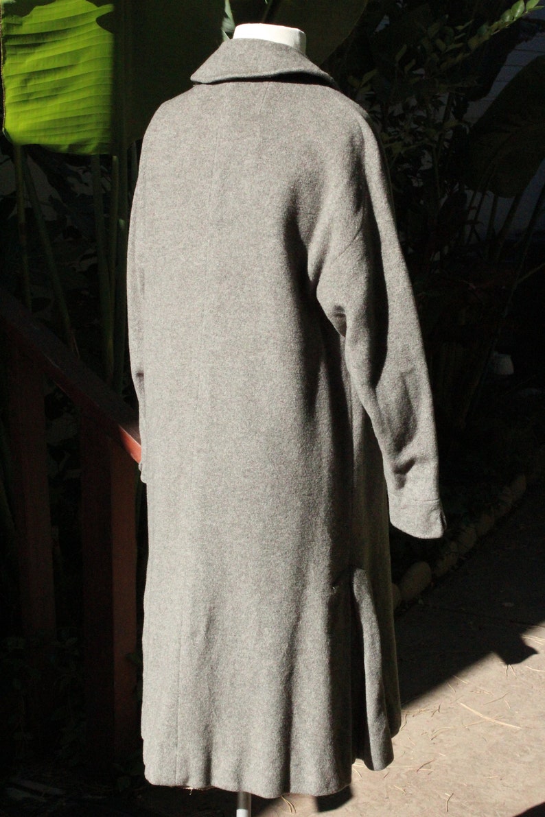 May include: A long, gray wool coat with a wide collar and long sleeves. The coat has a slight A-line shape and a split in the back for ease of movement. The coat is displayed on a white mannequin.