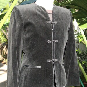 May include: A dark gray velvet jacket with a mandarin collar and decorative frog closures. The jacket has long sleeves, a tailored fit, and a small pocket detail. The jacket is displayed on a mannequin.