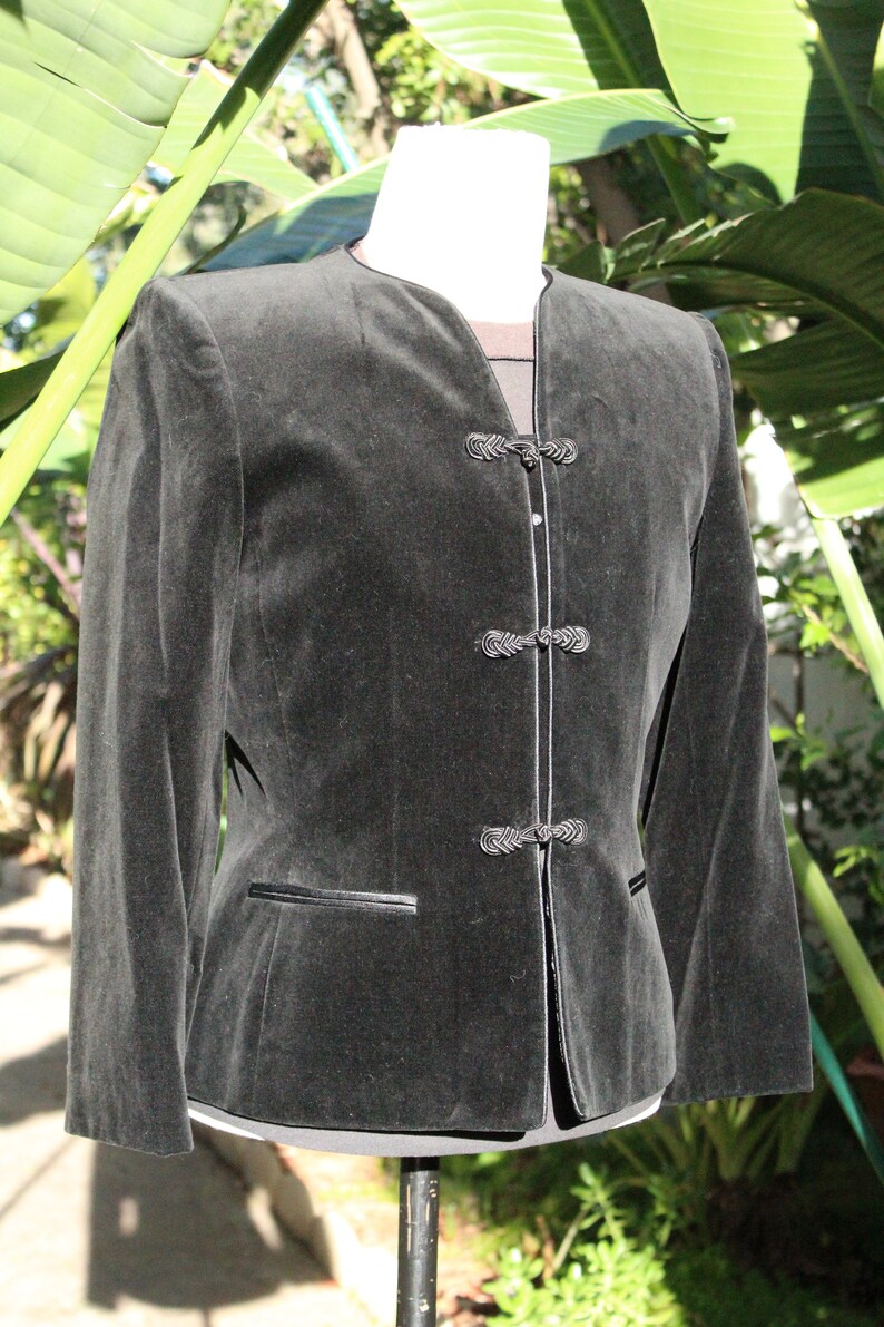 May include: A dark gray velvet jacket with a mandarin collar and decorative frog closures. The jacket has long sleeves, a tailored fit, and a small pocket detail. The jacket is displayed on a mannequin.