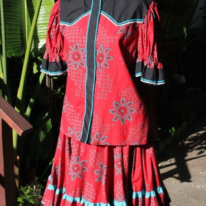 May include: A red two-piece outfit with a paisley pattern, black accents, and turquoise trim. The top has short sleeves and a round collar. The skirt has a ruffled hem. The outfit is displayed on a mannequin.