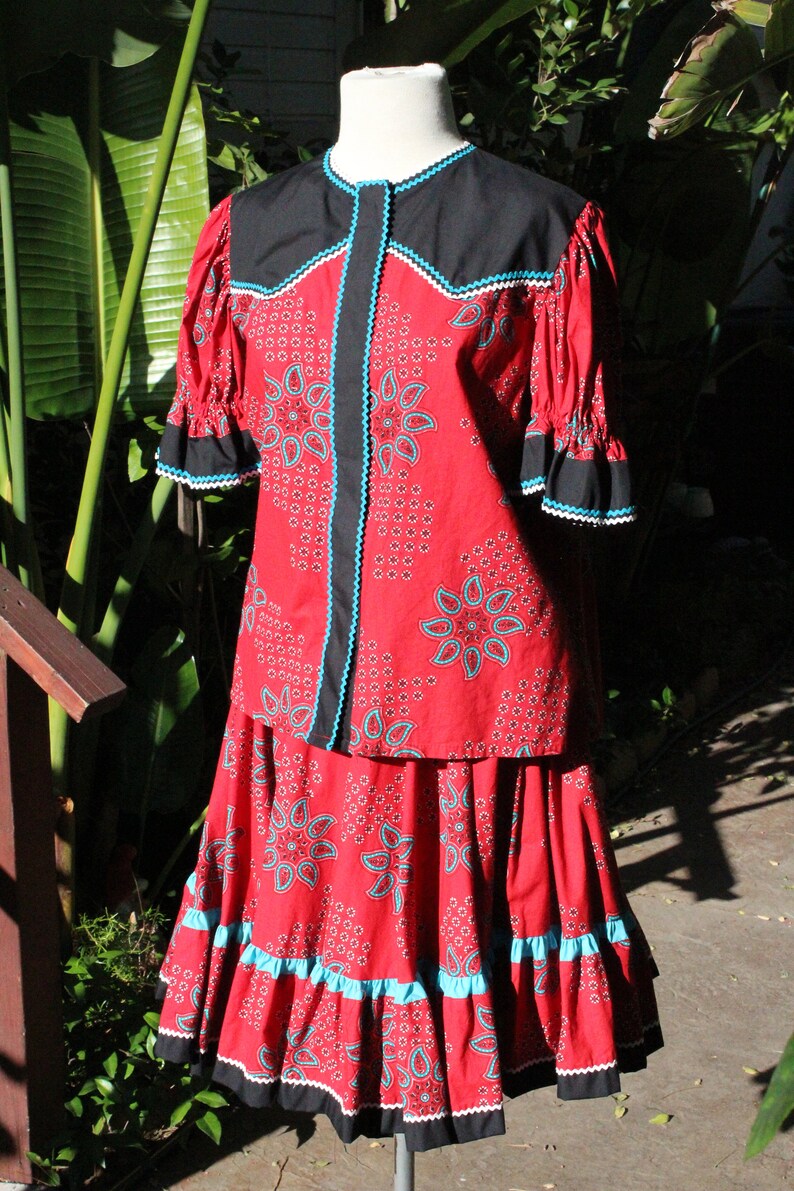 May include: A red two-piece outfit with a paisley pattern, black accents, and turquoise trim. The top has short sleeves and a round collar. The skirt has a ruffled hem. The outfit is displayed on a mannequin.
