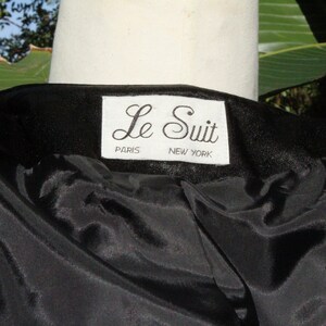 May include: Close-up of a black garment with a satin-like lining. The label reads "Le Suit PARIS NEW YORK" in a white rectangular tag. The garment is draped over a white mannequin, with green foliage in the background.