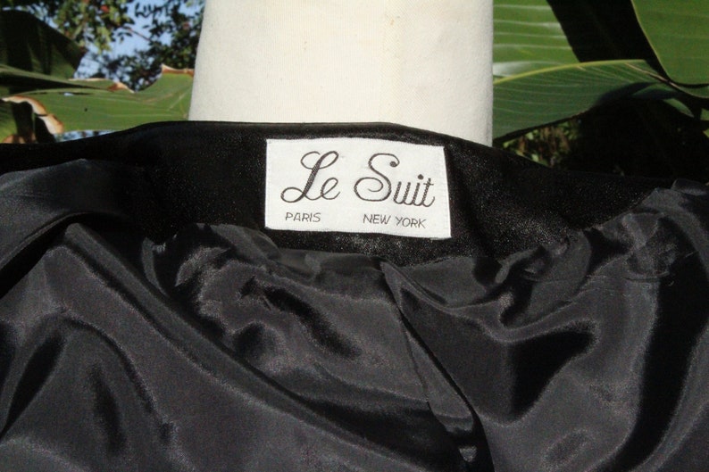 May include: Close-up of a black garment with a satin-like lining. The label reads "Le Suit PARIS NEW YORK" in a white rectangular tag. The garment is draped over a white mannequin, with green foliage in the background.