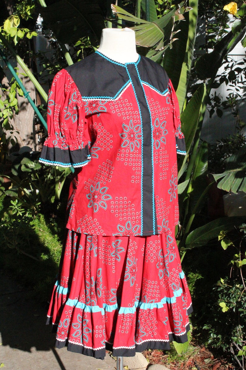 May include: A red two-piece outfit with a paisley pattern, black accents, and turquoise trim. The top has short sleeves and a black collar. The skirt has a ruffled hem. The outfit is displayed on a mannequin.
