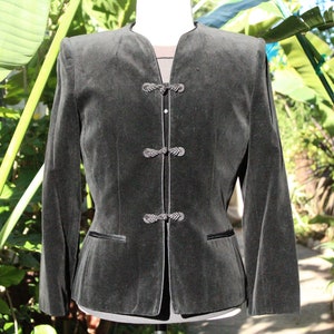 May include: A dark gray velvet jacket with a mandarin collar and three decorative frog closures. The jacket has long sleeves, a tailored fit, and two small pockets. The background features lush green foliage.