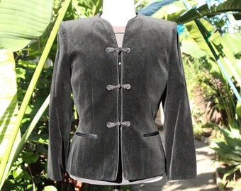 May include: A dark grey velvet jacket with a mandarin collar and three decorative frog closures. The jacket has long sleeves, a tailored fit, and two small pockets. The background features lush green foliage.