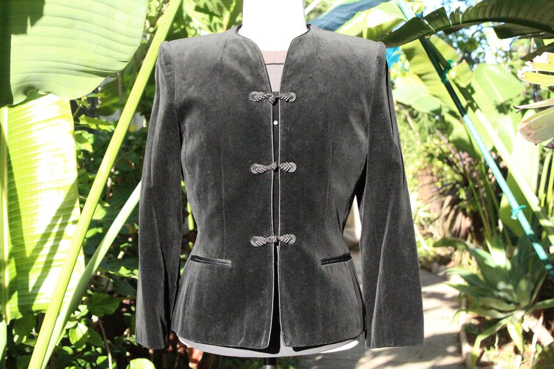 May include: A dark gray velvet jacket with a mandarin collar and three decorative frog closures. The jacket has long sleeves, a tailored fit, and two small pockets. The background features lush green foliage.