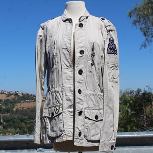 May include: A light beige jacket with multiple pockets, zippers, and black buttons. The jacket features patches with logos, including a "DAKAR" patch on the sleeve. The jacket is displayed on a mannequin against a backdrop of a blue sky and greenery.