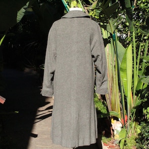 May include: A long, gray wool coat with a classic design. The coat features a notched collar, long sleeves, and a knee-length hem. The back view shows the coat's tailored silhouette, perfect for cooler weather. The coat is displayed outdoors with lush greenery in the background.
