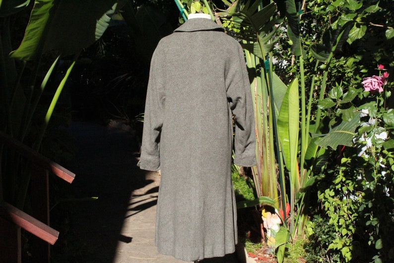 May include: A long, gray wool coat with a classic design. The coat features a notched collar, long sleeves, and a knee-length hem. The back view shows the coat's tailored silhouette, perfect for cooler weather. The coat is displayed outdoors with lush greenery in the background.