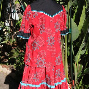 May include: A red two-piece outfit with a floral pattern and turquoise accents. The top has a black yoke and sleeves with black and white trim. The skirt has a ruffled hem with turquoise trim. The outfit is displayed on a mannequin.