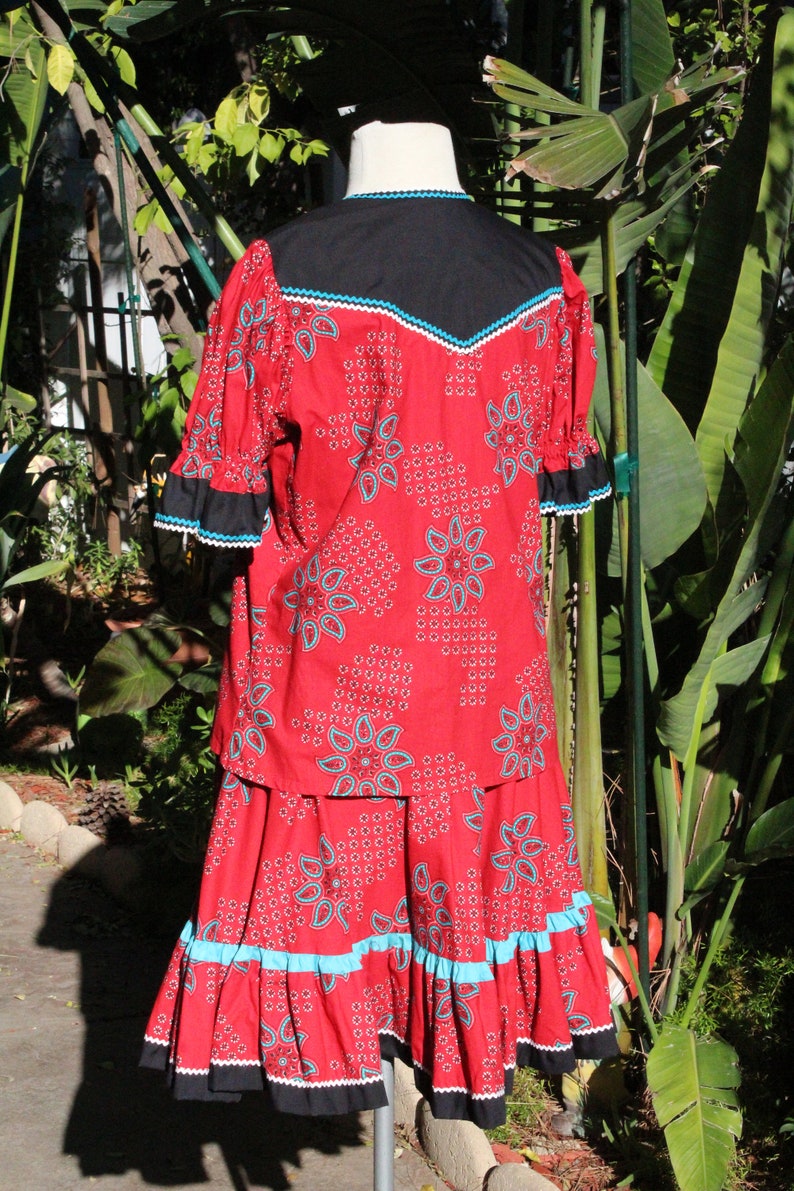 May include: A red two-piece outfit with a floral pattern and turquoise accents. The top has a black yoke and sleeves with black and white trim. The skirt has a ruffled hem with turquoise trim. The outfit is displayed on a mannequin.