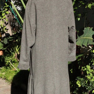 May include: A gray wool coat with a notched collar and long sleeves. The coat is knee-length and has a classic, tailored design. The coat is displayed on a mannequin in front of a green leafy background.