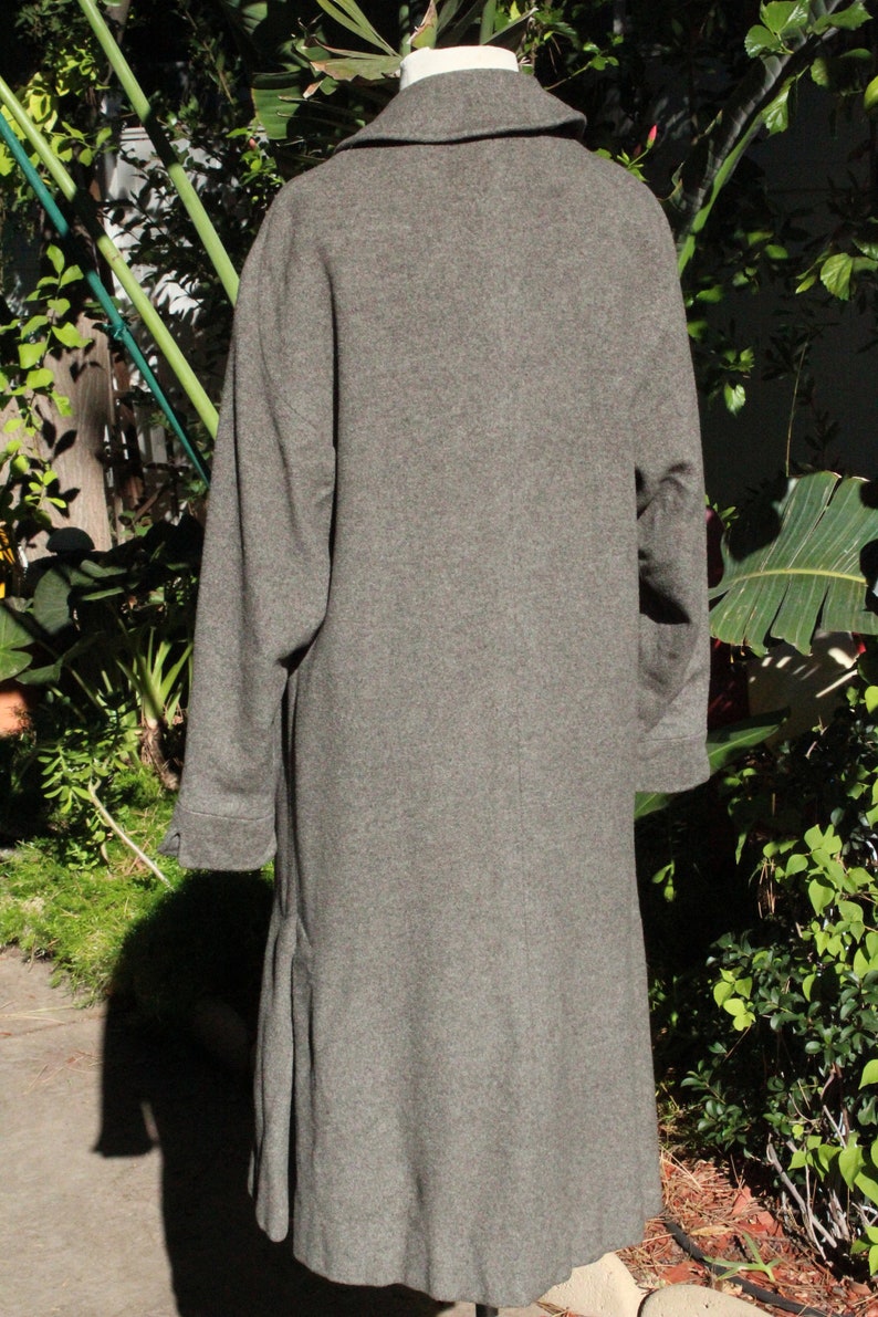 May include: A gray wool coat with a notched collar and long sleeves. The coat is knee-length and has a classic, tailored design. The coat is displayed on a mannequin in front of a green leafy background.